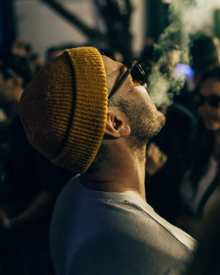 A man wearing a yellow beanie and sunglasses tilts his head back as smoke drifts upward.