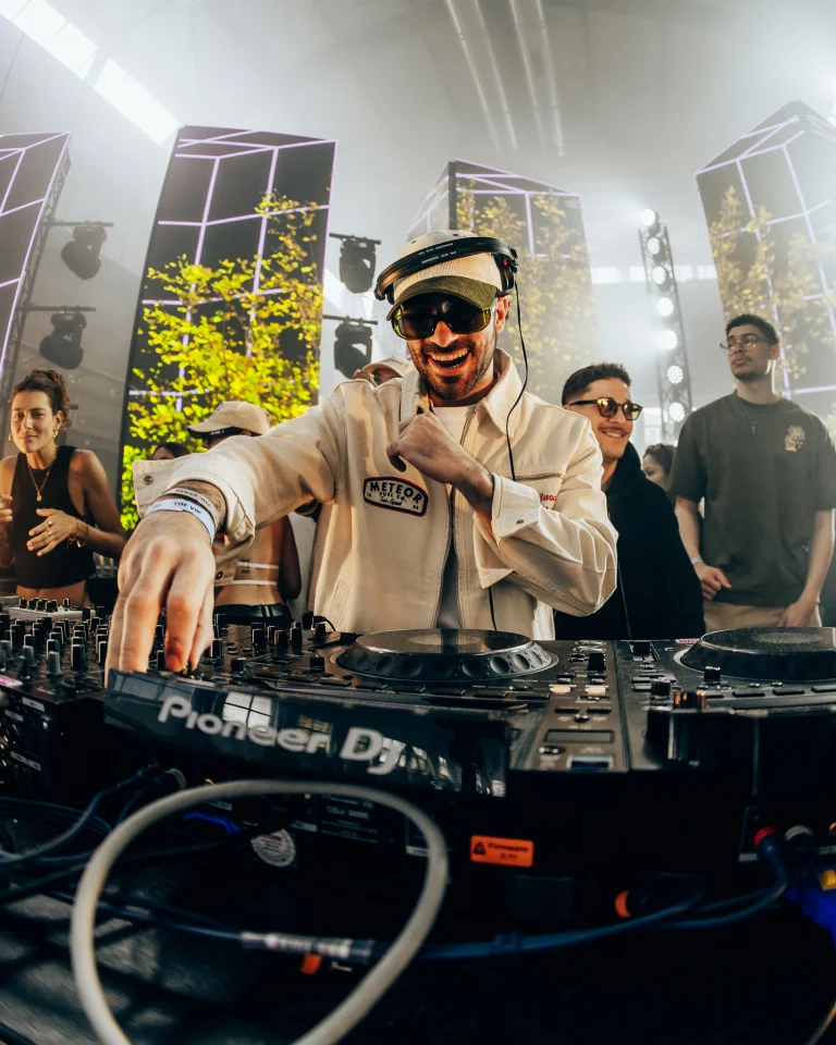 A smiling man wearing a cap and sunglasses operates a Pioneer DJ mixing console, surrounded by other people in a bright indoor venue.
