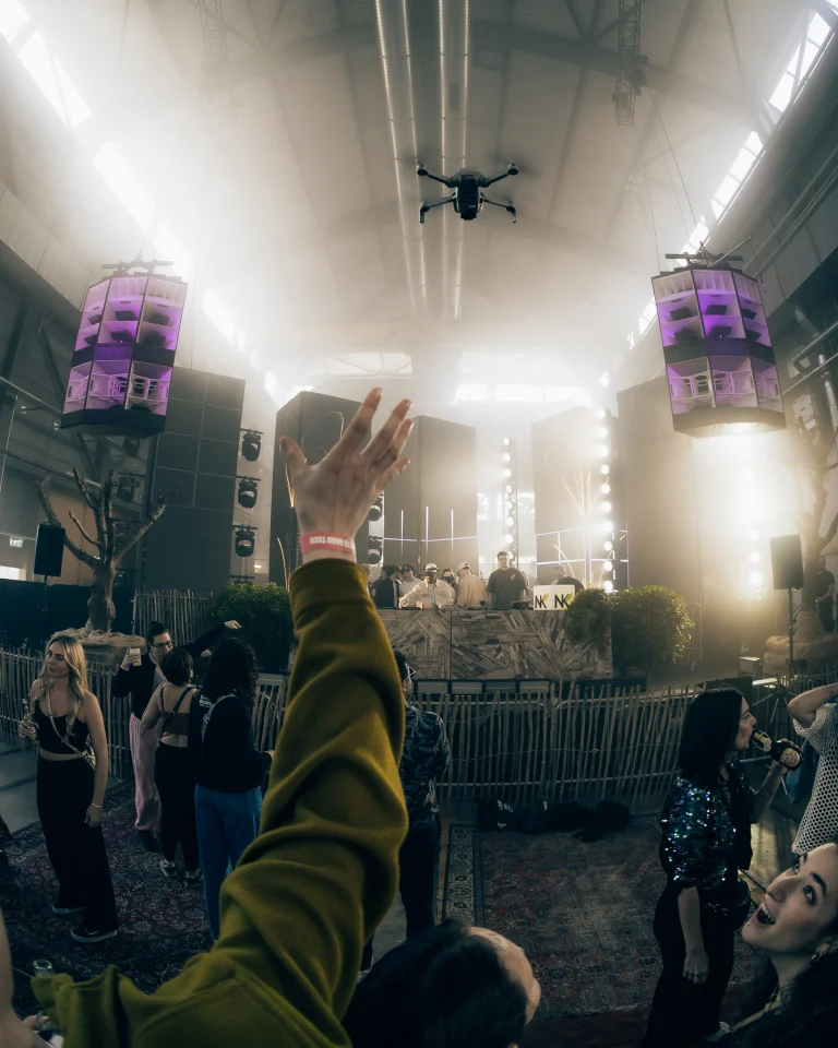 A drone flies overhead in a large industrial hall filled with hazy light, while a person in the foreground raises a hand.