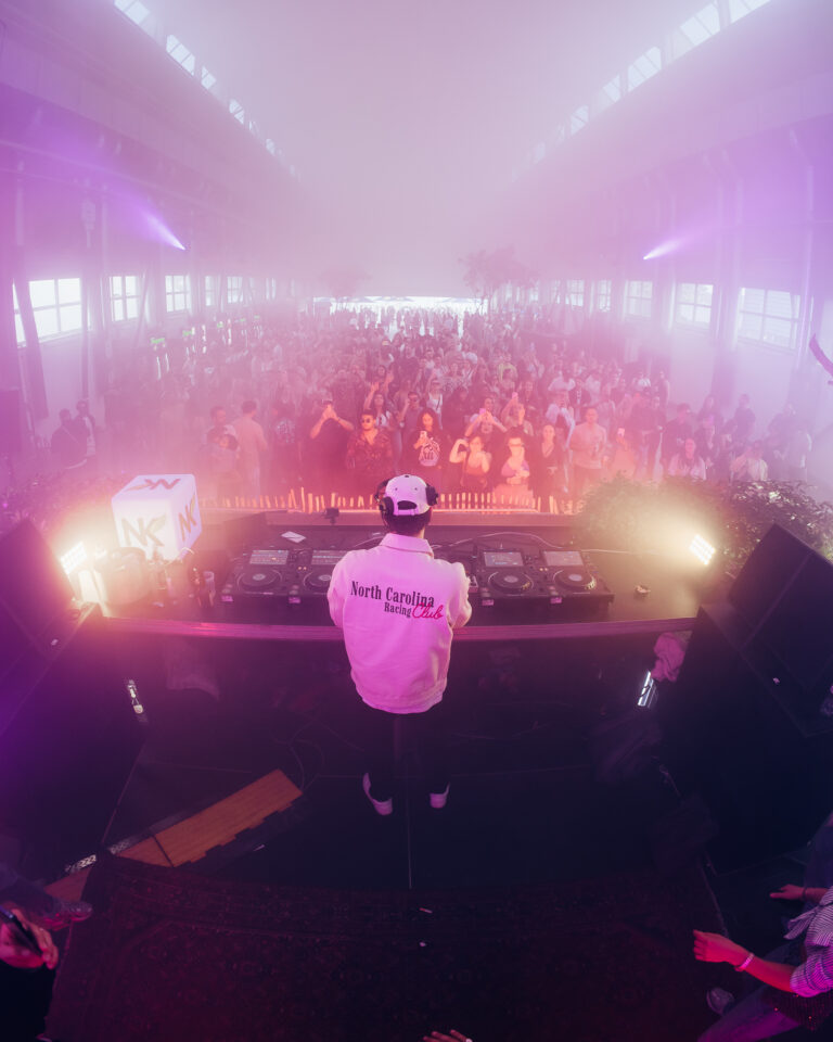 Ein Mann steht hinter einem DJ-Pult in einer nebligen Halle mit violettem Licht und blickt auf eine grosse Menge.
