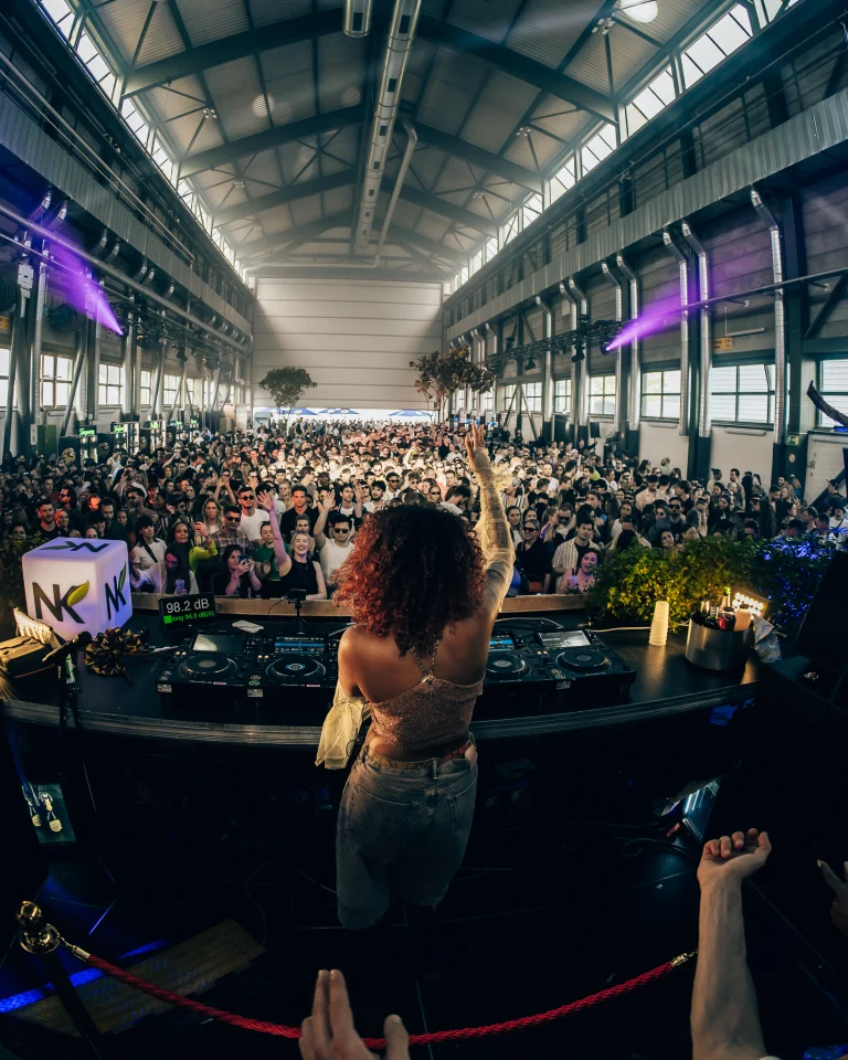 A woman stands with her back to the camera at a DJ booth, raising her hand before a large crowd in a spacious industrial hall.