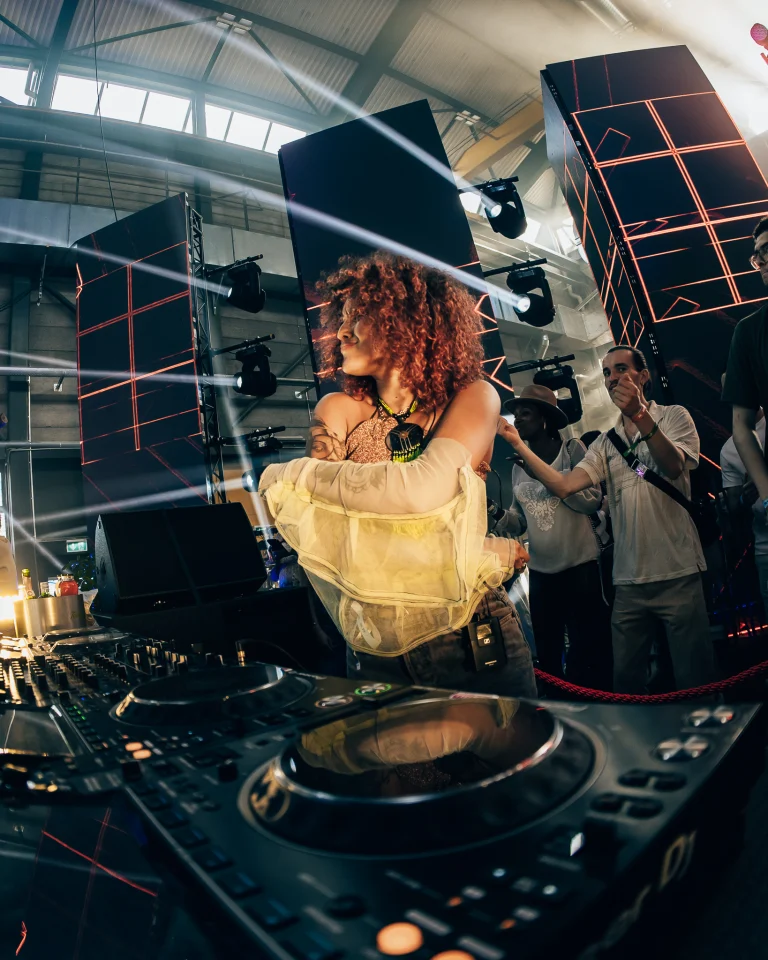 A woman with curly reddish hair stands behind a DJ mixer in an industrial venue, with people visible in the background.