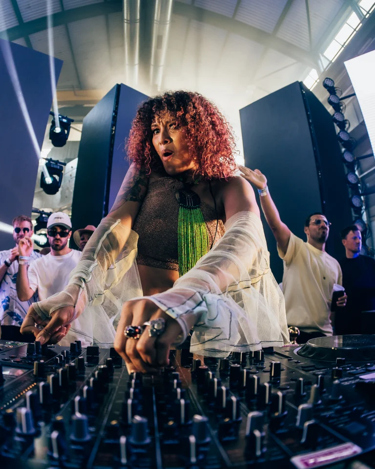 A woman with curly red hair performs at a DJ mixing console under stage lights, flanked by men in the background.