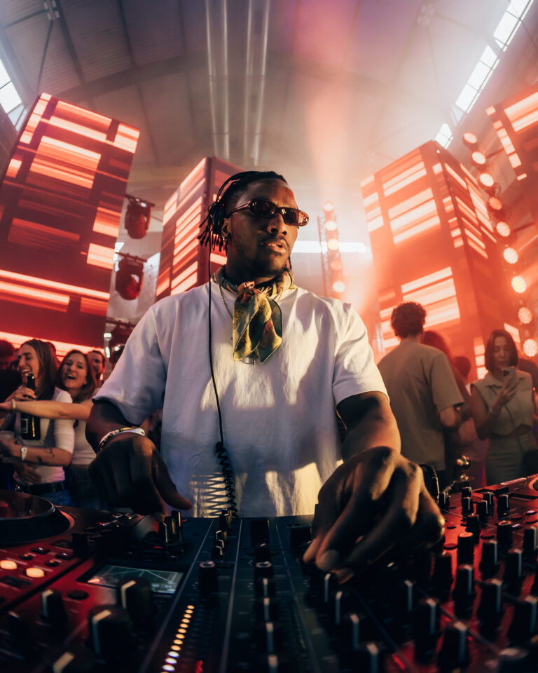 Ein Mann mit Sonnenbrille und gemustertem Schal steht unter heller, orangefarbenem Bühnenbeleuchtung an einem DJ-Mixer, umgeben von einer Menschenmenge.