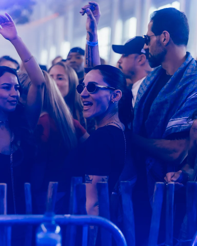 A smiling woman with sunglasses and a raised arm stands in a crowded crowd, bathed in blue light.