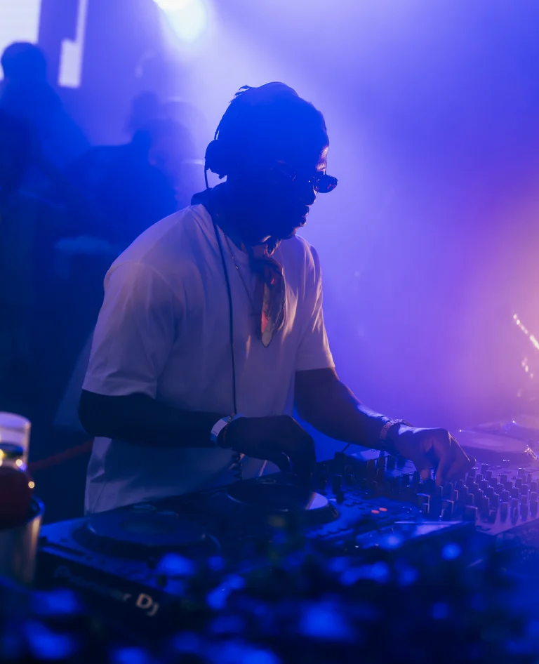 A man wearing headphones and sunglasses stands behind a mixing console, illuminated by blue and purple stage lights.