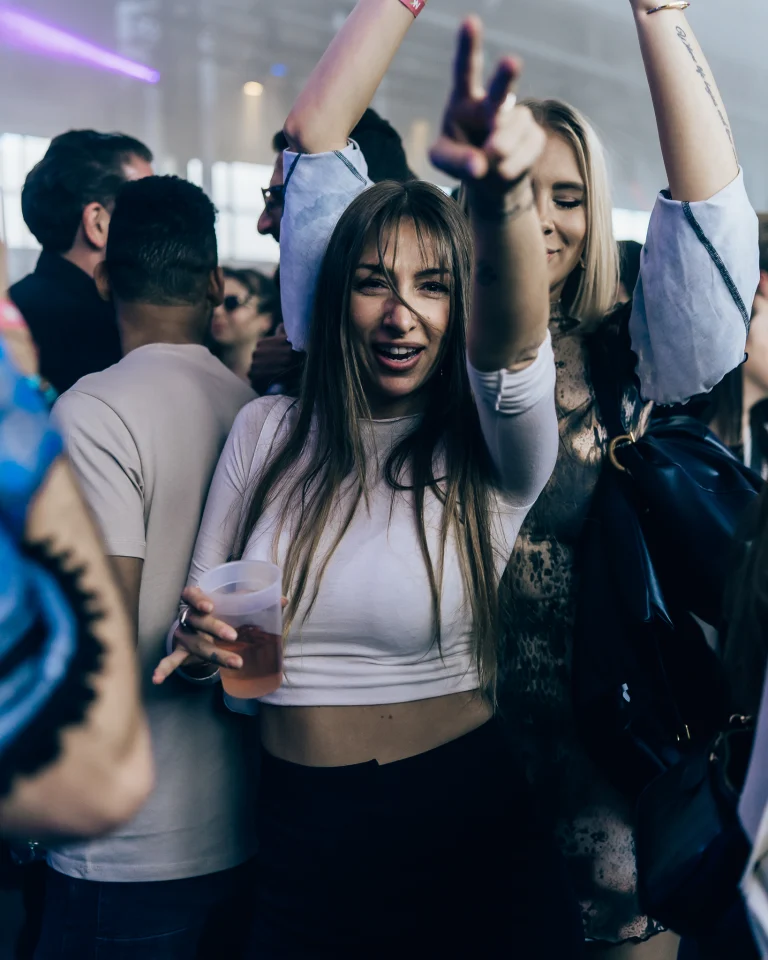 A woman holding a plastic cup raises her hand in a peace sign while dancing among a crowd indoors.