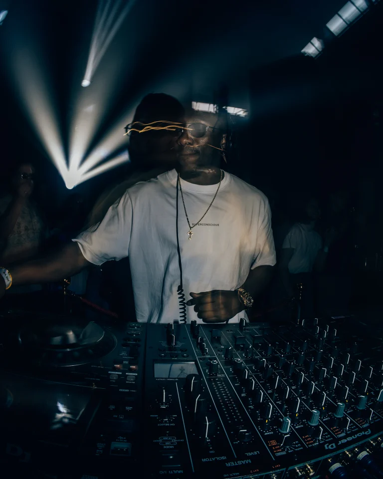 A double exposure shot of a man in a white t-shirt operating a Pioneer DJ mixer in a dimly lit club with a bright light beam in the background.