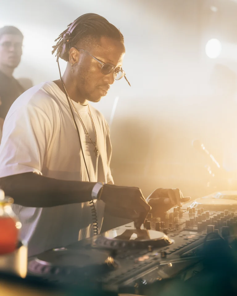A man wearing sunglasses and headphones stands behind a mixing console, operating the equipment under warm stage lighting.