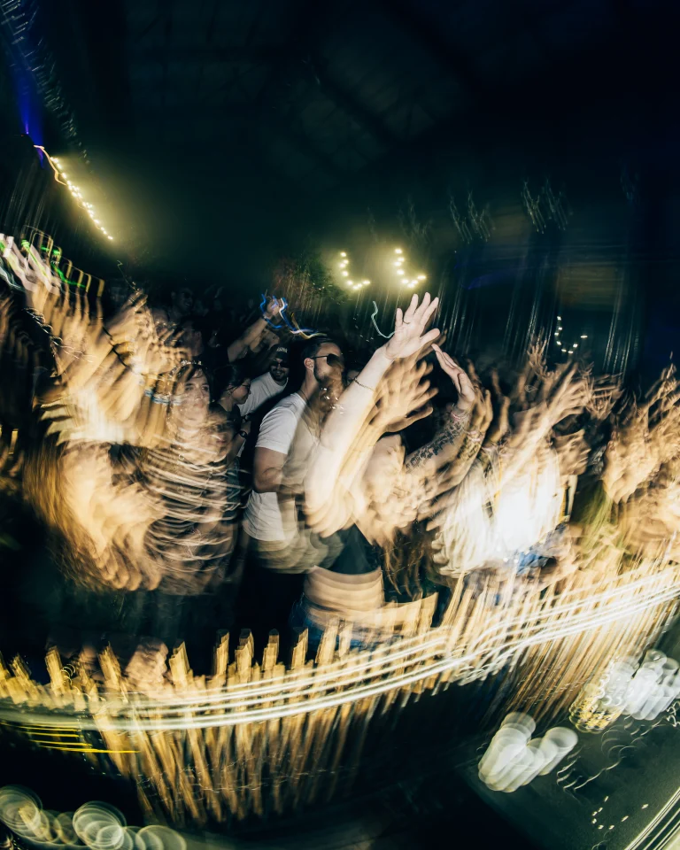 A crowd of people with blurred motion effects raises their hands in a dimly lit venue.