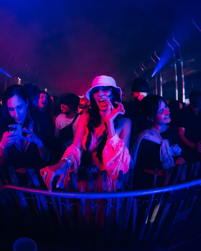 A woman wearing a white bucket hat stands behind a barrier in a crowd, illuminated by blue and pink lights.