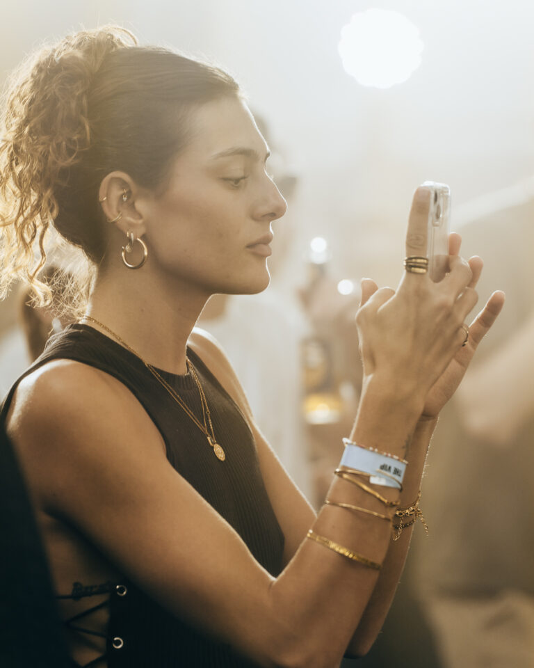 Eine Frau im Profil hält ihr Smartphone mit beiden Händen hoch, um ein Foto zu machen, und trägt mehrere goldene Armbänder, Ringe und Ohrringe.