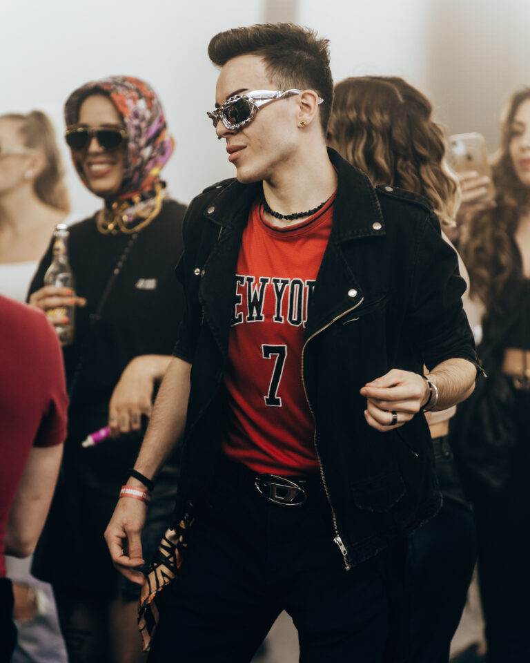 Ein Mann mit futuristischer Sonnenbrille, schwarzer Jacke und rotem Shirt mit der Aufschrift NEW YORK 7 steht zwischen Menschen.