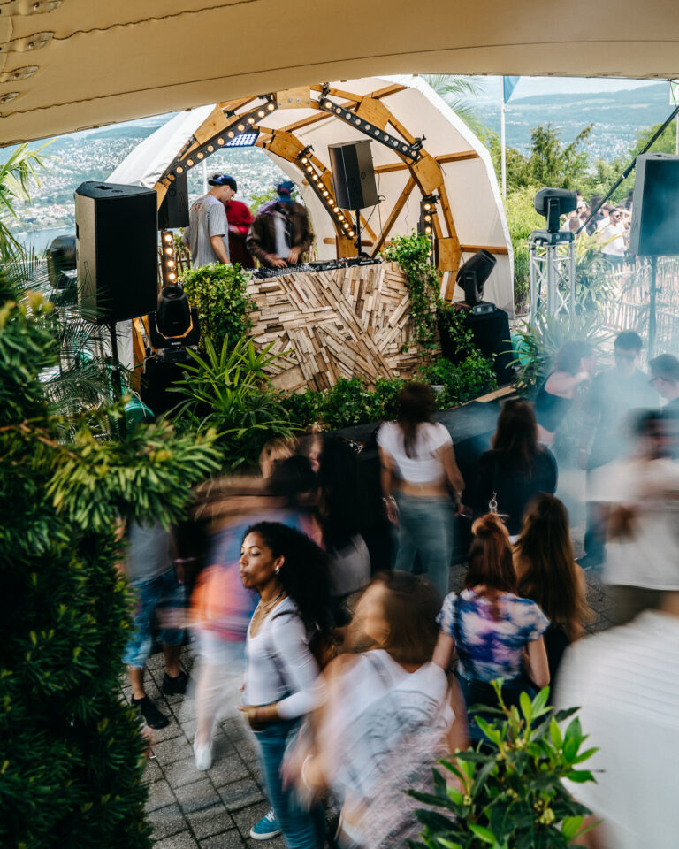 Eine Weitaufnahme einer Open-Air-Tagesparty unter einem weißen Zelt, bei der im Vordergrund Männer und Frauen tanzen, während DJs auf einer holzverkleideten Bühne mit Lautsprechern und Lichtanlagen auf eine ferne Stadtlandschaft blicken.