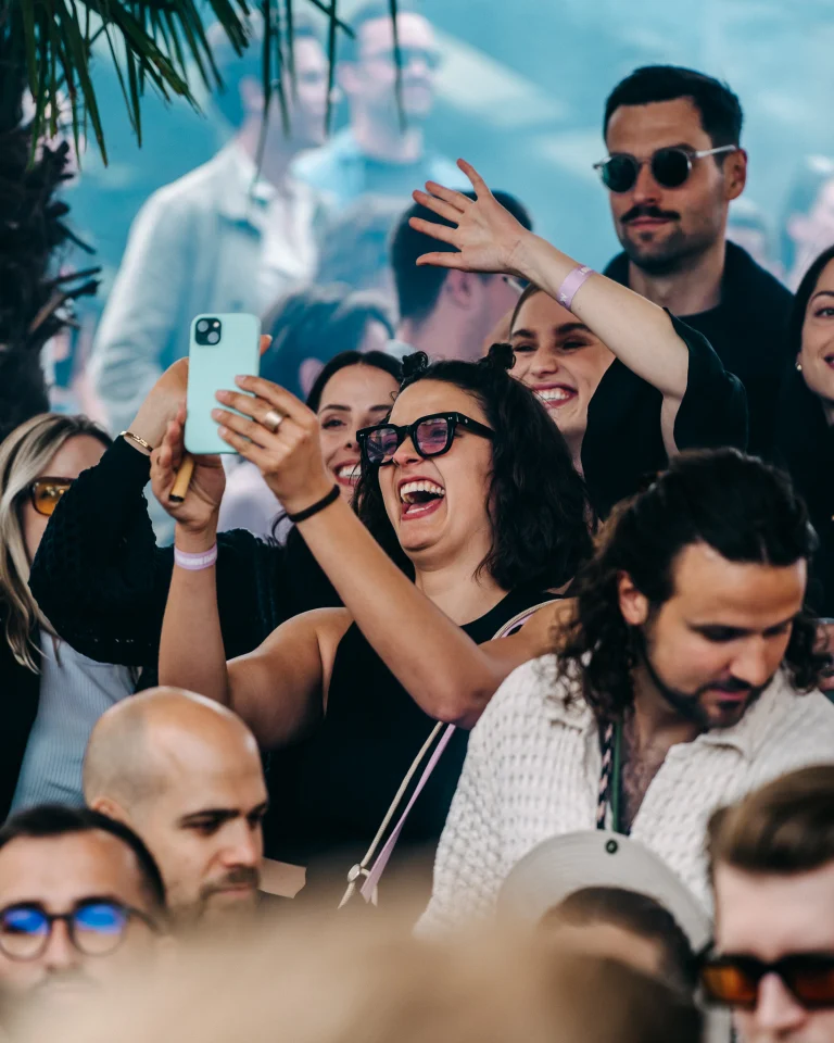 A smiling woman in glasses holds up a phone to take a photo among a crowd of people.