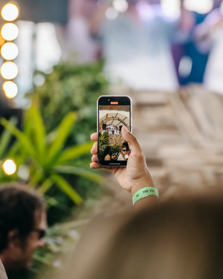 A hand holding a smartphone up to record video, with a green wristband bearing the text NK THE VIP on the wrist.