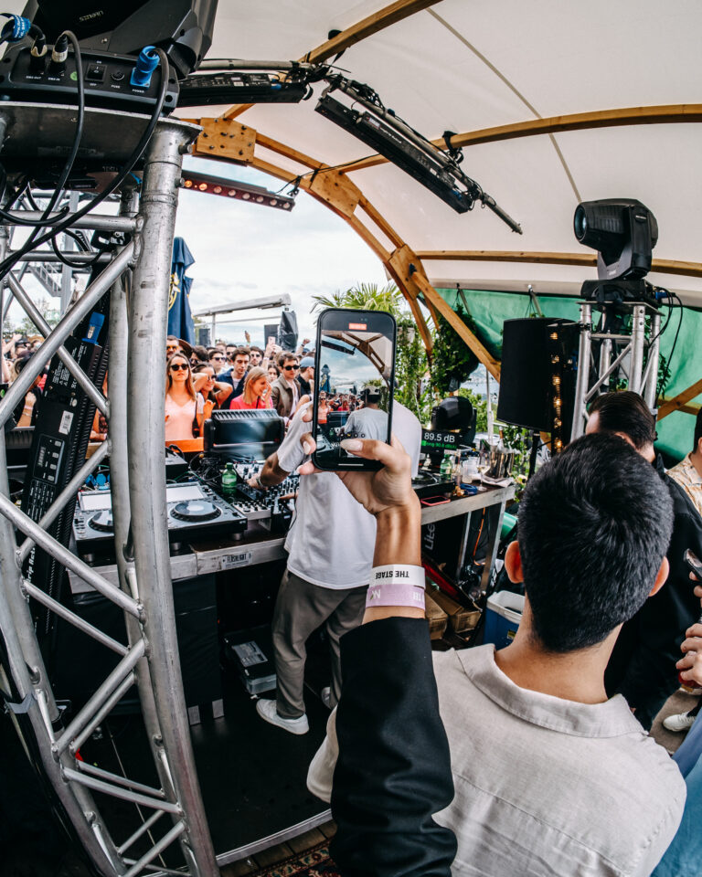 Eine Frau hält ein Smartphone hoch, um ein DJ-Pult von hinten durch einen Mann im Vordergrund unter einem Festzelt mit Bühnenkonstruktion aufzunehmen.