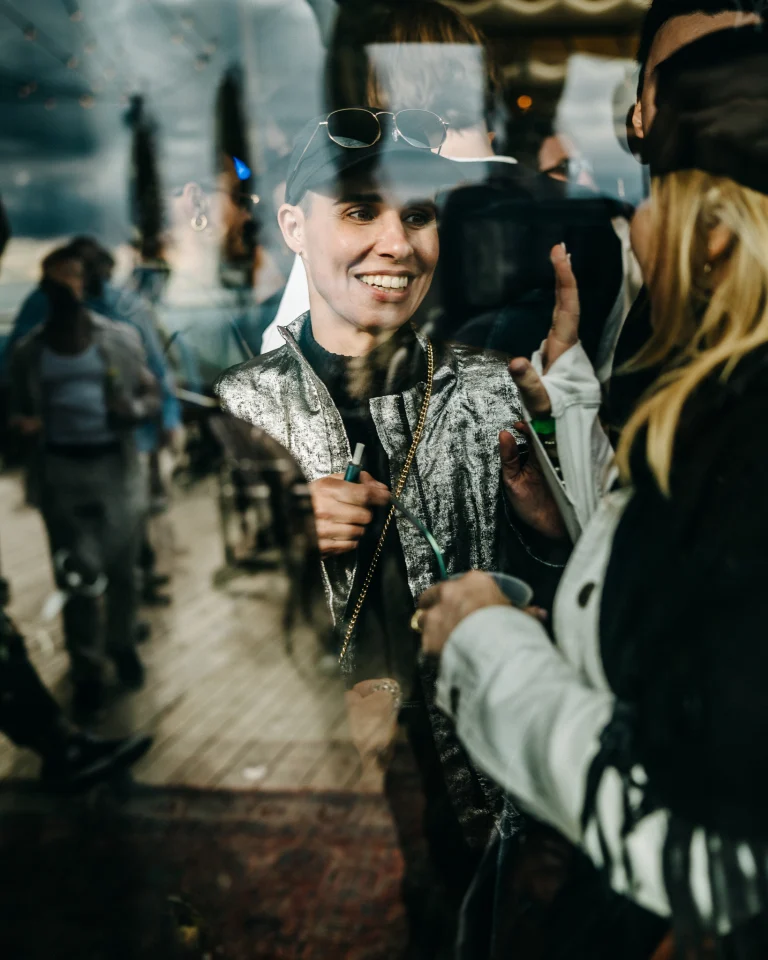 A smiling person wearing a shiny silver jacket and a cap looks toward a woman in profile, partially obscured by reflections on glass.