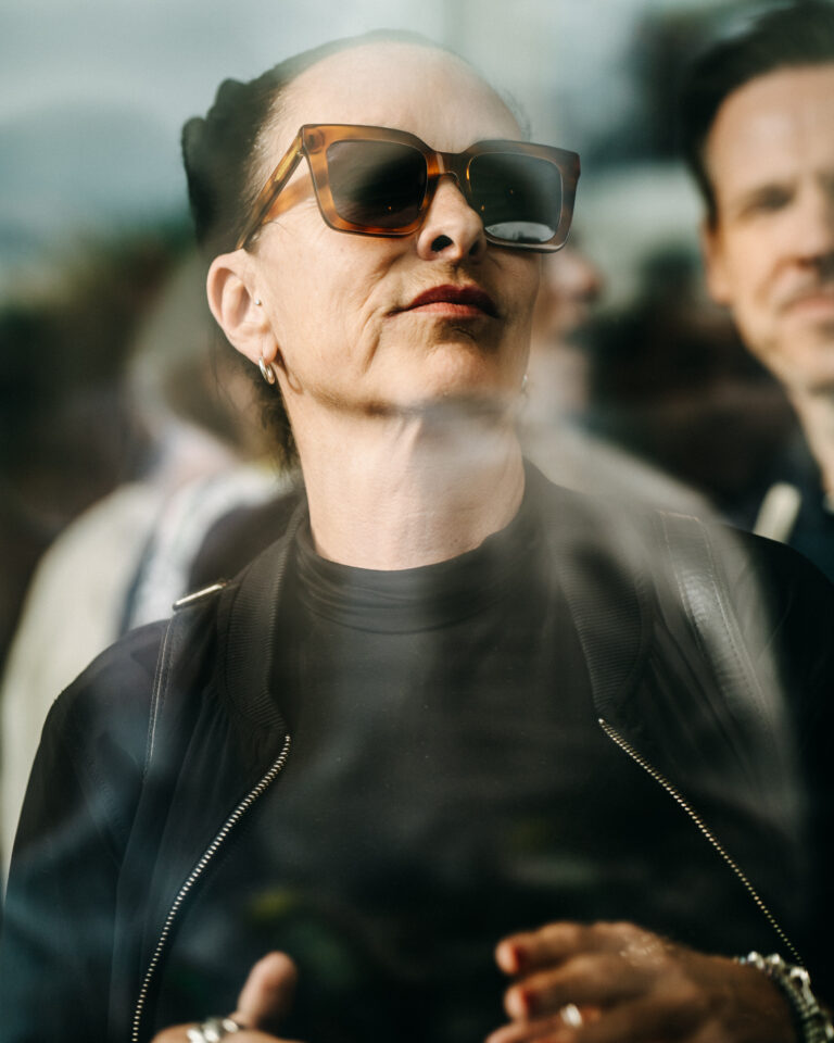 Eine Frau mit großer Sonnenbrille und dunkler Jacke mit Reißverschluss, teilweise verdeckt durch eine spiegelnde Oberfläche, mit einem Mann im Hintergrund.