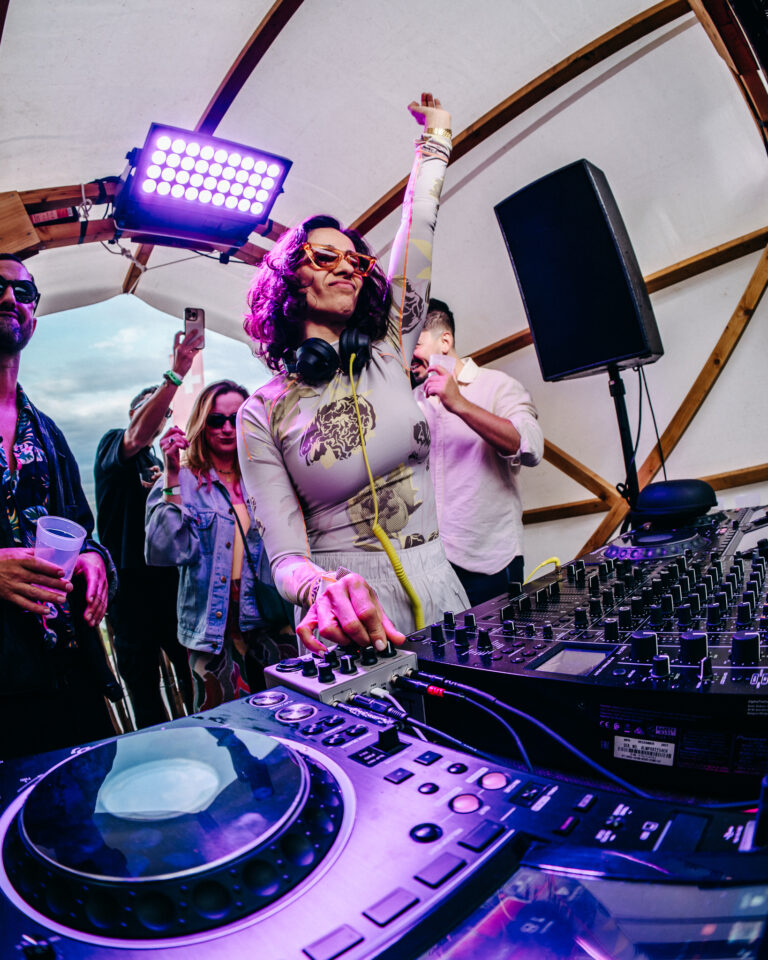 Eine Frau steht unter einem Zelt hinter einem DJ-Mischpult, reckt einen Arm in die Höhe und bedient unter violettem Licht die Geräte.
