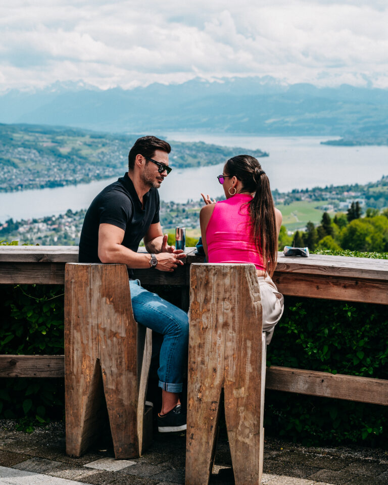 Ein Mann und eine Frau sitzen an einem hohen Holztisch mit Blick auf eine See- und Berglandschaft.
