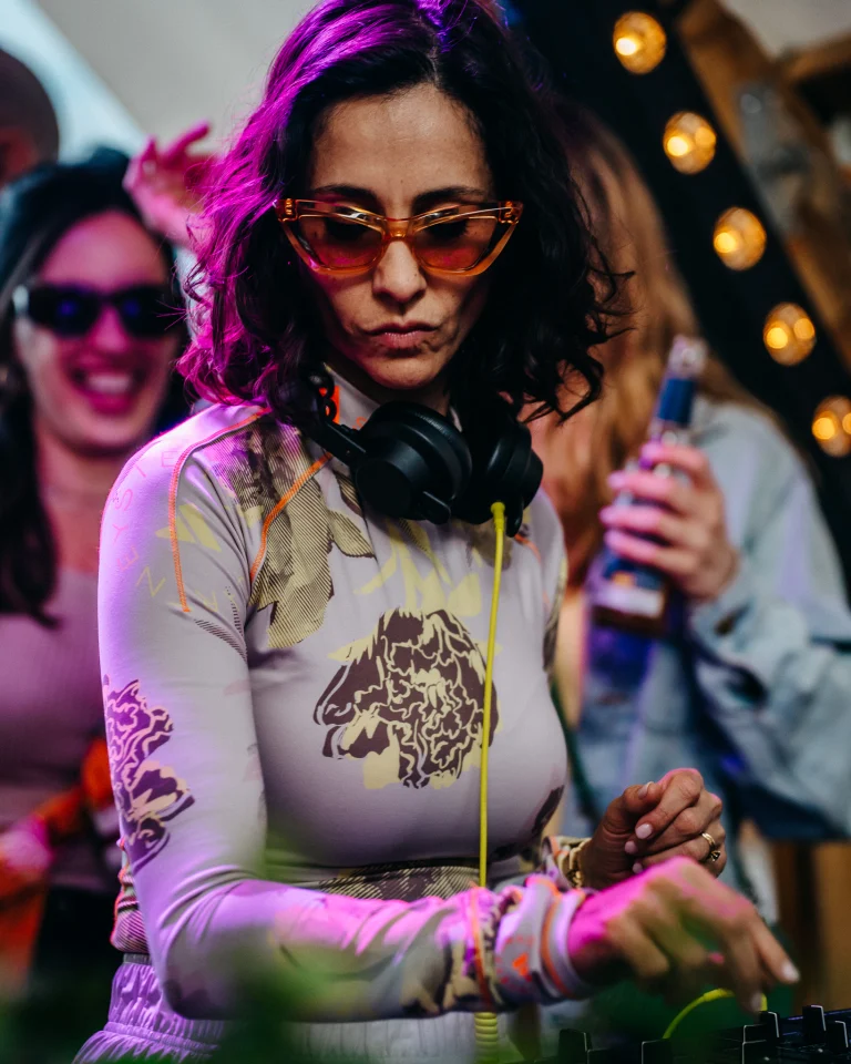 A woman wearing orange-tinted cat-eye sunglasses and headphones stands at a mixing console under warm stage lighting.