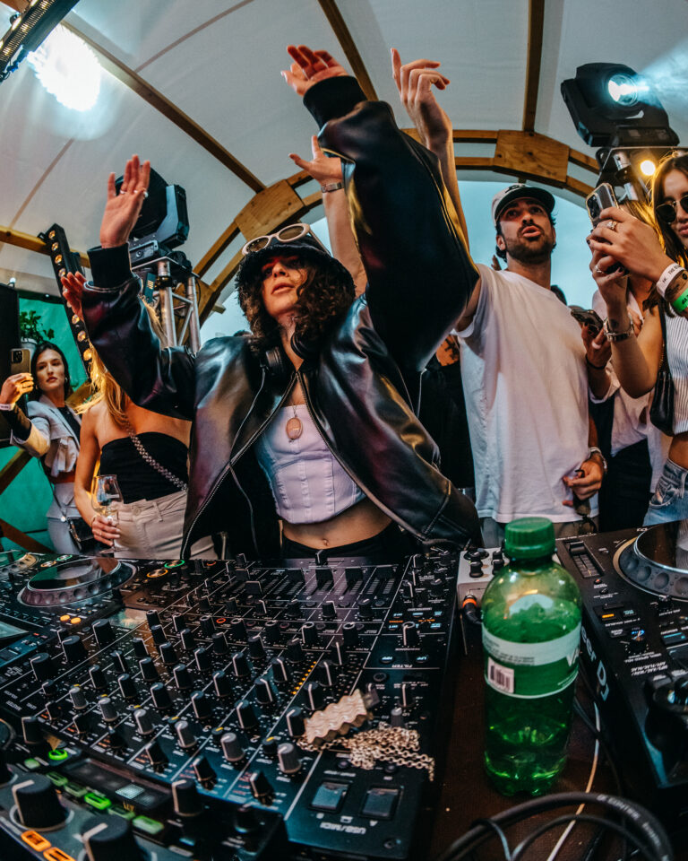 Eine Frau in einer schwarzen Jacke und mit Hut hebt die Arme, während sie in einer vollen Location hinter einem DJ-Mixer steht, aufgenommen mit einem Fisheye-Objektiv.