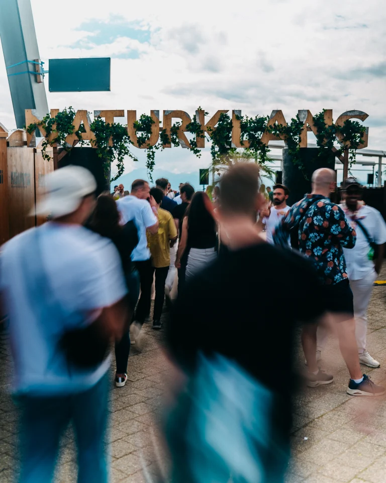 People walking through an entrance archway decorated with foliage and the sign NATURKLANG under an overcast sky.