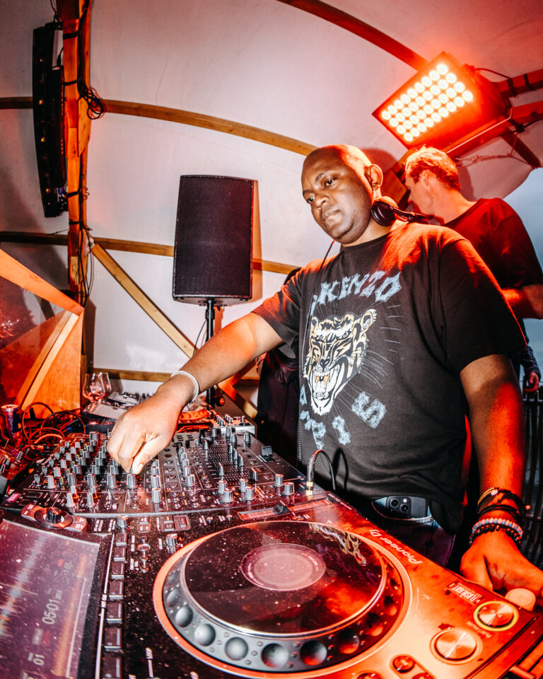 Ein Mann steht unter warmer Bühnenbeleuchtung hinter einem DJ-Mischpult und verstellt Regler, während ein anderer Mann im Hintergrund steht.