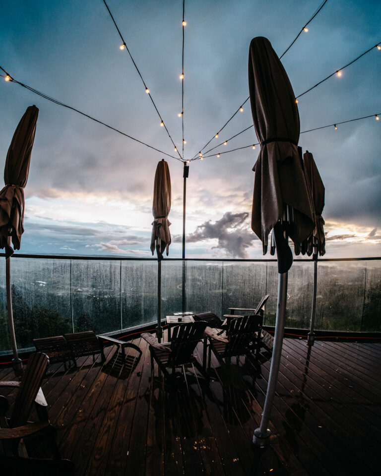 Überdachte Holzterrasse mit geschlossenen Sonnenschirmen und Stühlen unter Lichterketten vor bewölktem Abendhimmel.