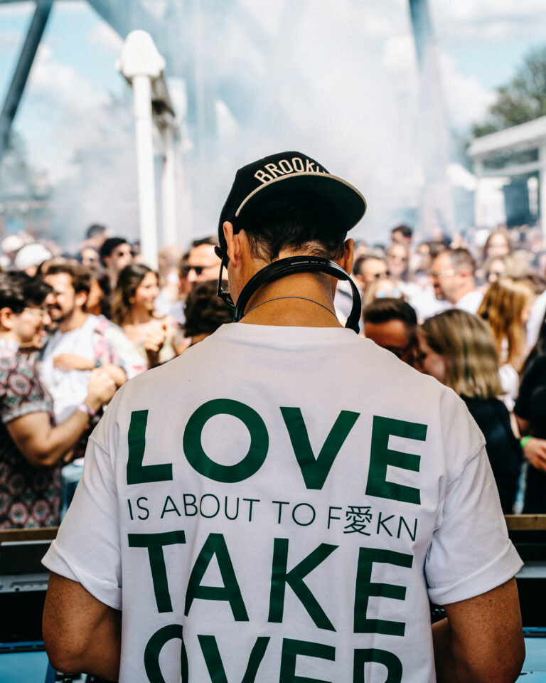 Ein Mann mit einem weißen T-Shirt und grüner Aufschrift LOVE IS ABOUT TO F*KN TAKE OVER steht im Tageslicht vor einer Menschenmenge.