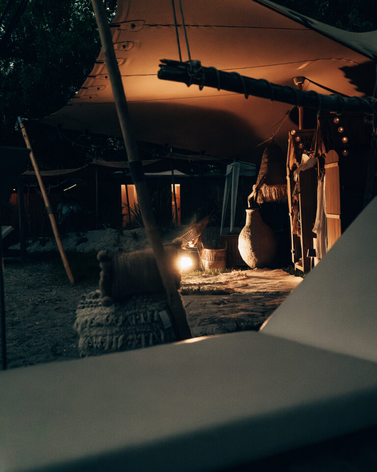 Ein beleuchtetes Zeltlager bei Nacht mit warmem Licht auf einem großen Krug, strukturierten Kissen und Holzstrukturen.