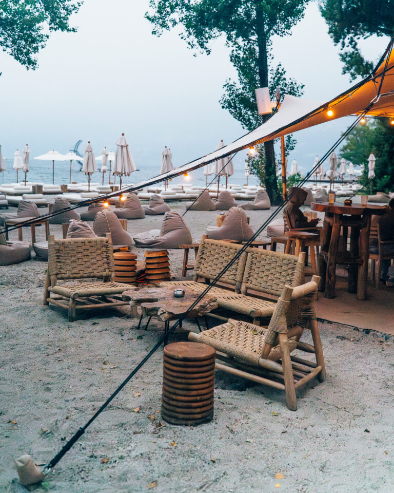 Strandlounge im Freien mit geflochtenen Holzsesseln, niedrigen Tischen und Sitzsäcken unter einem Baldachin mit Lichterkette in der Dämmerung.