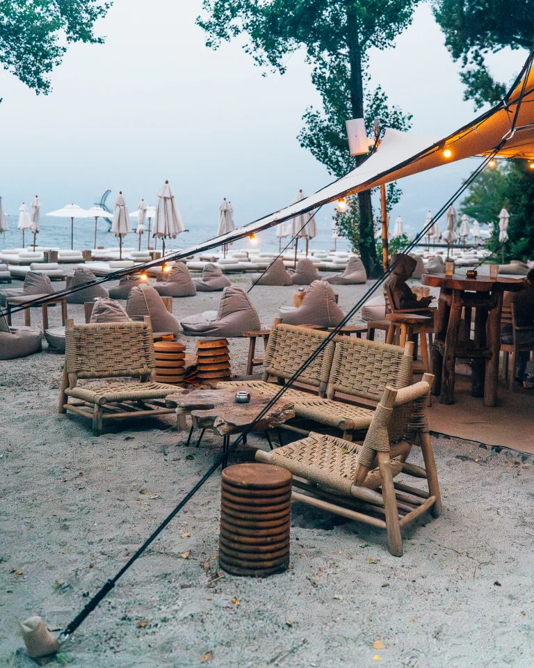 Outdoor beach lounge with woven wooden armchairs, low tables, and beanbags under a canopy string-lit at dusk.