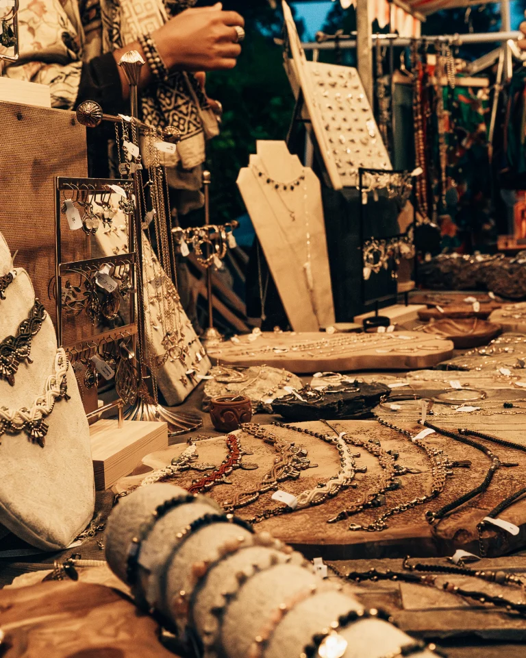 A market stall displays an assortment of necklaces, bracelets, and earrings arranged on stands and wooden trays, with a person's hands visible in the background.