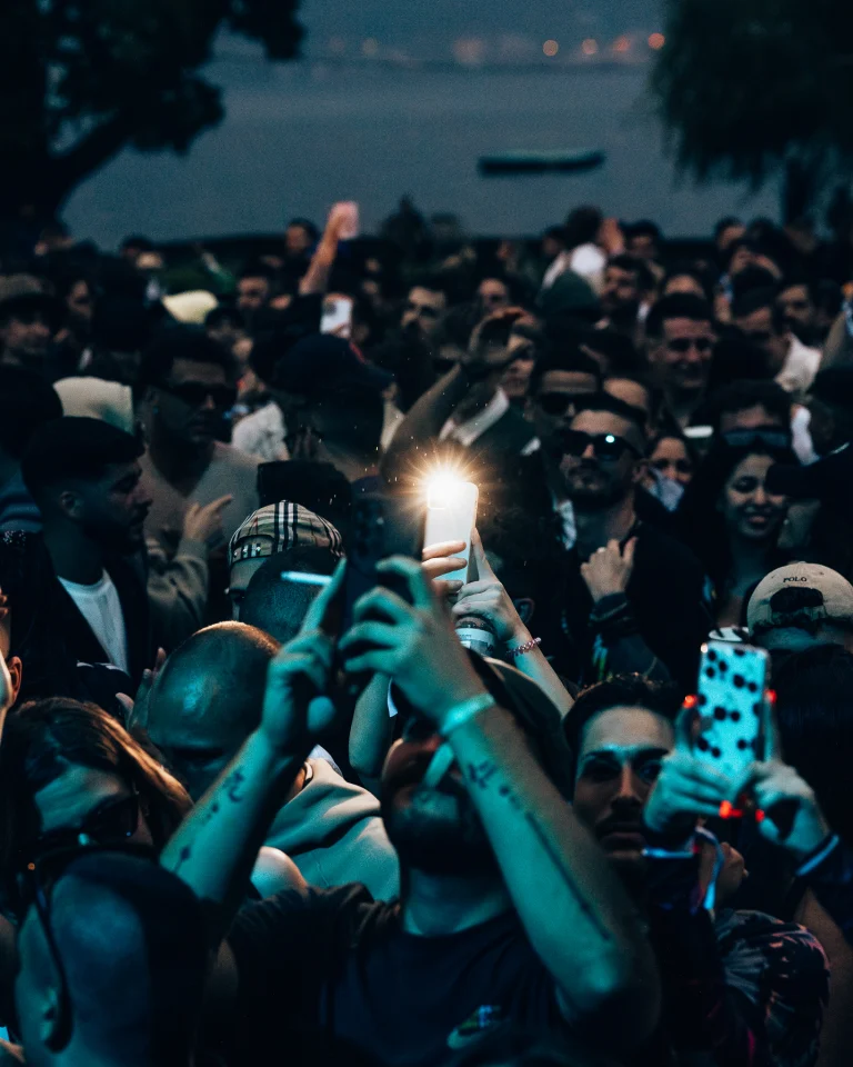 A dense crowd of people at night holding up smartphones to take photos or record videos under cool blue lighting, with a bright flash illuminating the center.
