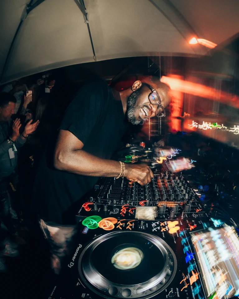 A man wearing glasses leans over a DJ mixing console in a dimly lit venue, with motion-blurred lights reflecting in the foreground.
