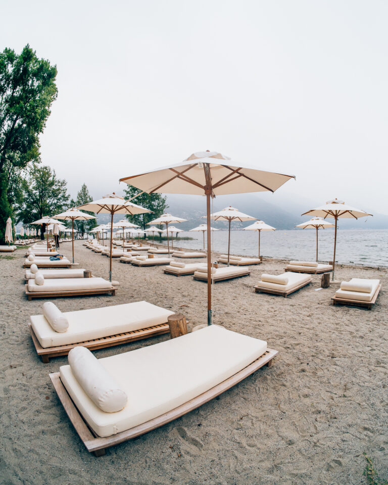 Reihen von beige farbenen Liegen und Sonnenschirmen säumen einen Sandstrand unter bedecktem Himmel an einem ruhigen See.
