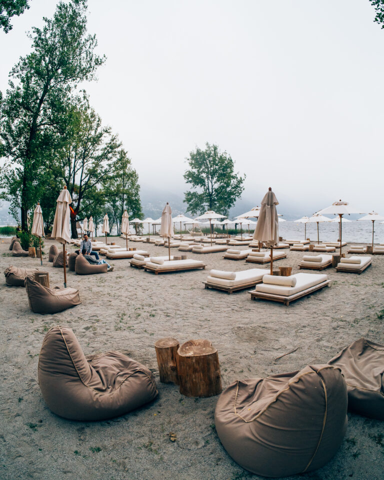 Ein bewölkter Strand am See zeigt Reihen weißer Liegen mit geschlossenen Sonnenschirmen und verstreute Sitzsäcke auf Sand unter Bäumen.