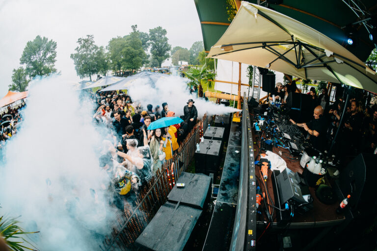 Eine Weitwinkel-Aufnahme aus der Vogelperspektive zeigt ein volles Open-Air-Konzert, bei dem Rauch über einer dichten Menschenmenge aufsteigt, während rechts ein Mann an einem DJ-Pult unter einem großen Schirm auflegt.