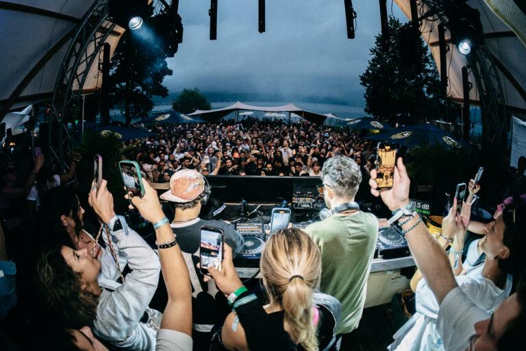 DJs stehen hinter einem Mischpult und blicken auf eine große Menschenmenge vor einer Open-Air-Bühne am Wasser, während Besucher Smartphones in die Höhe halten.