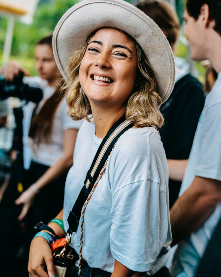 A smiling woman wearing a light-colored wide-brimmed hat and a white t-shirt looks back over her shoulder outdoors, with other people blurred in the background.