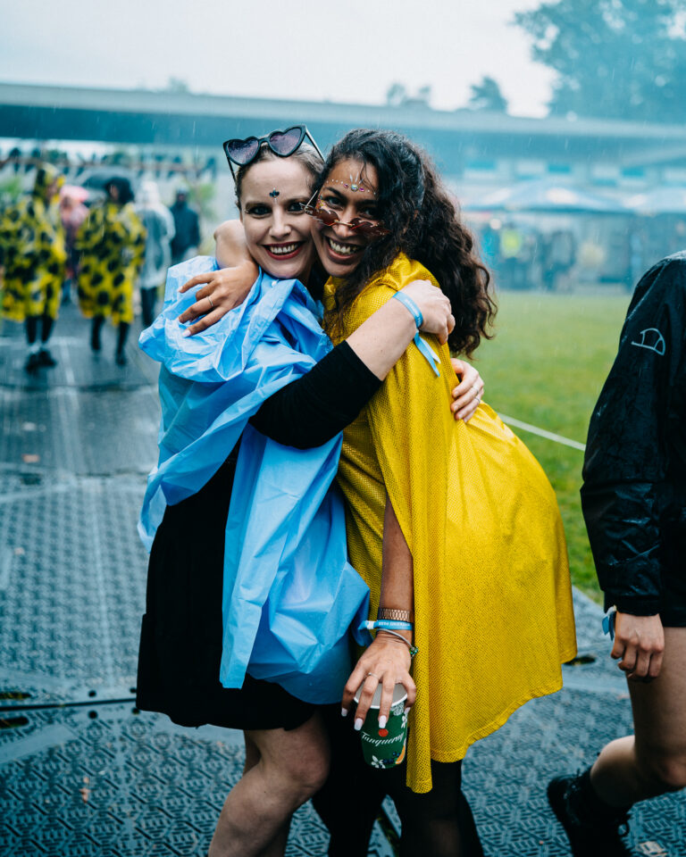 Zwei lächelnde Frauen in Regenmänteln umarmen sich im Freien, die eine in einem blauen Poncho, die andere in einem gelben Umhang.