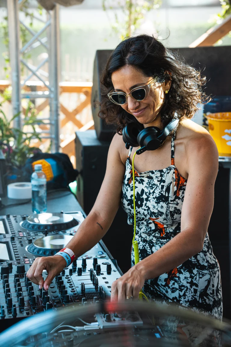 A woman wearing sunglasses and headphones stands at a DJ mixer, operating the controls outdoors in natural light.