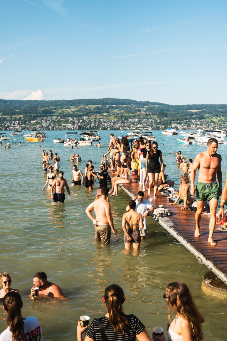 Ein gut gefüllter Holzsteg und flaches Wasser an einem sonnigen Tag, mit zahlreichen badenden und sich entspannenden Menschen nahe Booten auf einem großen See.