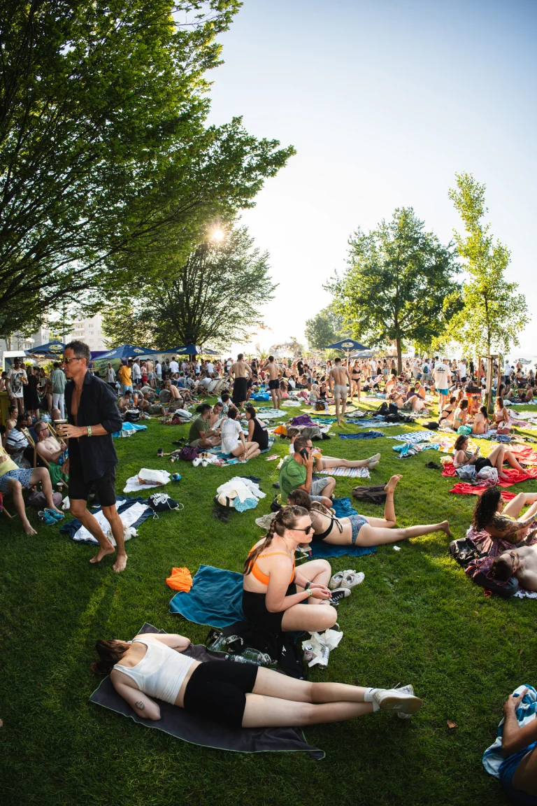 Fisheye view of a large crowd relaxing on a grassy park under bright sunlight, with a man walking barefoot in the foreground.