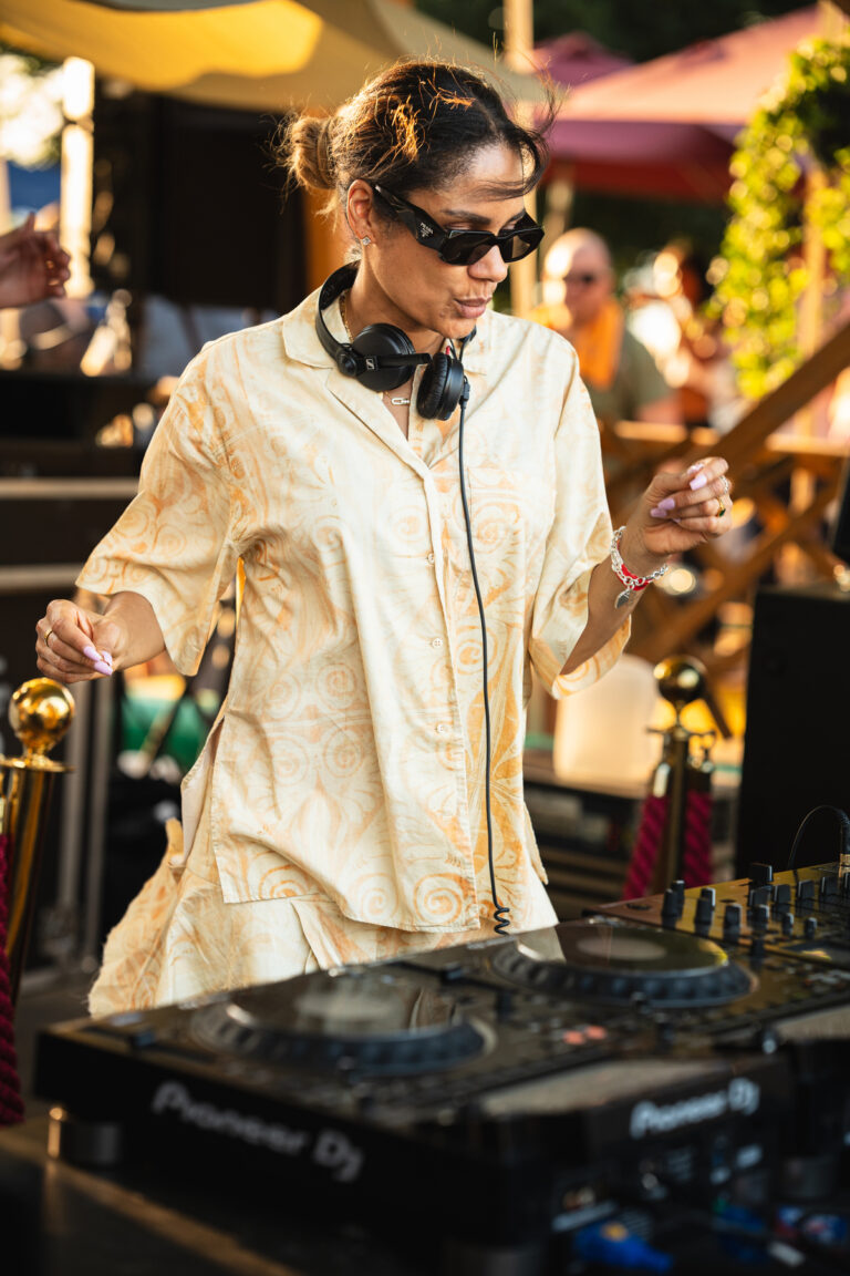Eine Frau mit Sonnenbrille und Kopfhörern steht im Freien hinter einem Pioneer-DJ-Pult und tanzt mit erhobenen Armen im warmen natürlichen Sonnenlicht.