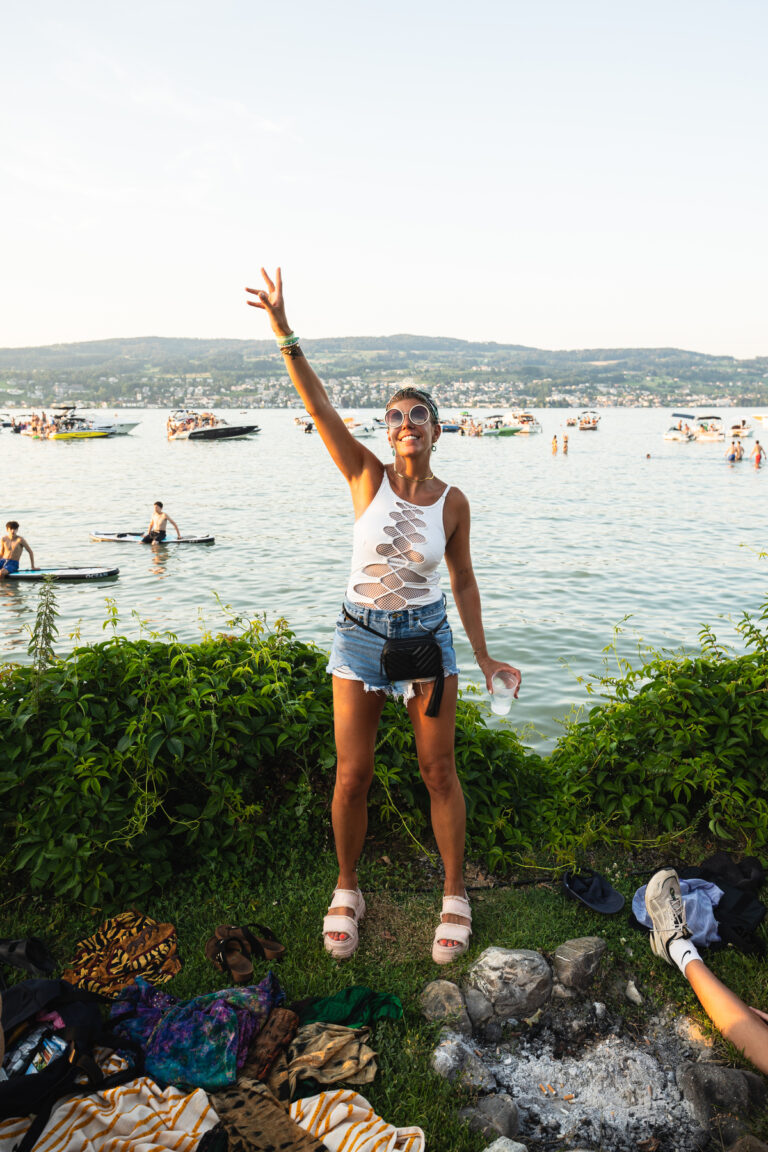 Eine Frau steht an einem See, hebt eine Hand zum Friedenszeichen und trägt eine Sonnenbrille sowie ein weißes Oberteil mit Ausschnitten, während im Hintergrund Boote auf dem Wasser fahren.