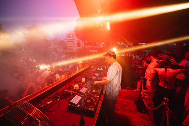 Ein Mann steht unter hellen Bühnenlichtern hinter einem DJ-Mischpult und legt bei einem Open-Air-Festival für eine dichte Menge auf.