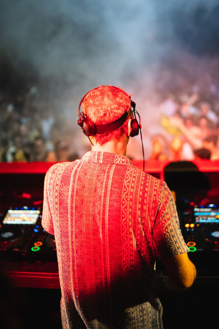 A man seen from behind wearing headphones and a patterned red short-sleeve shirt stands at a mixing console facing a blurred crowd under red lighting.
